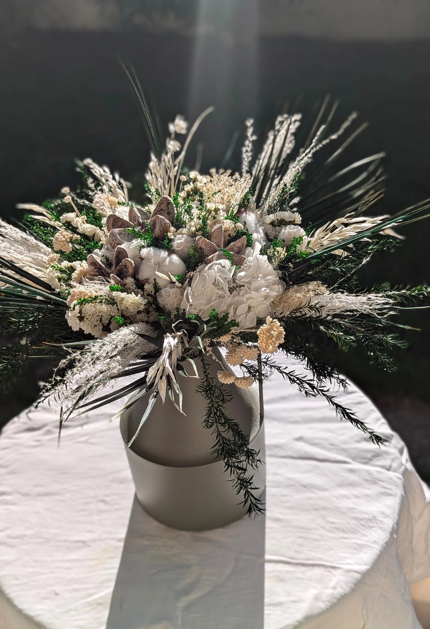 Composition en fleurs élégantes préservées mêlant achillée, coton, hortensias blancs et feuillages verts. Une création raffinée et durable, parfaite pour une ambiance hivernale et chaleureuse. Vase en métal vert  olive. A la lumière du jour. Bridgette