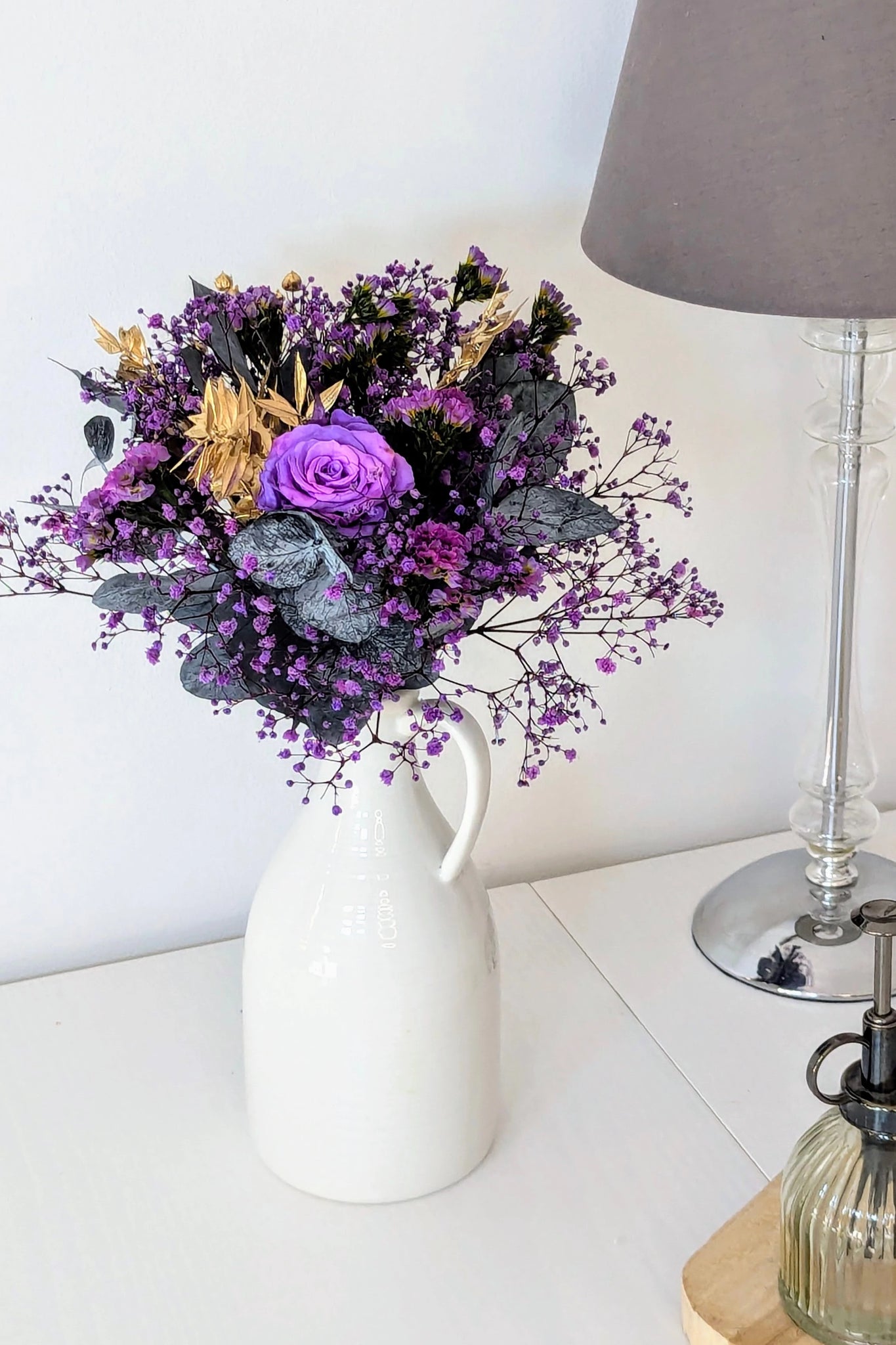 Bouquet violet et doré dans pichet blanc sur table blanche
