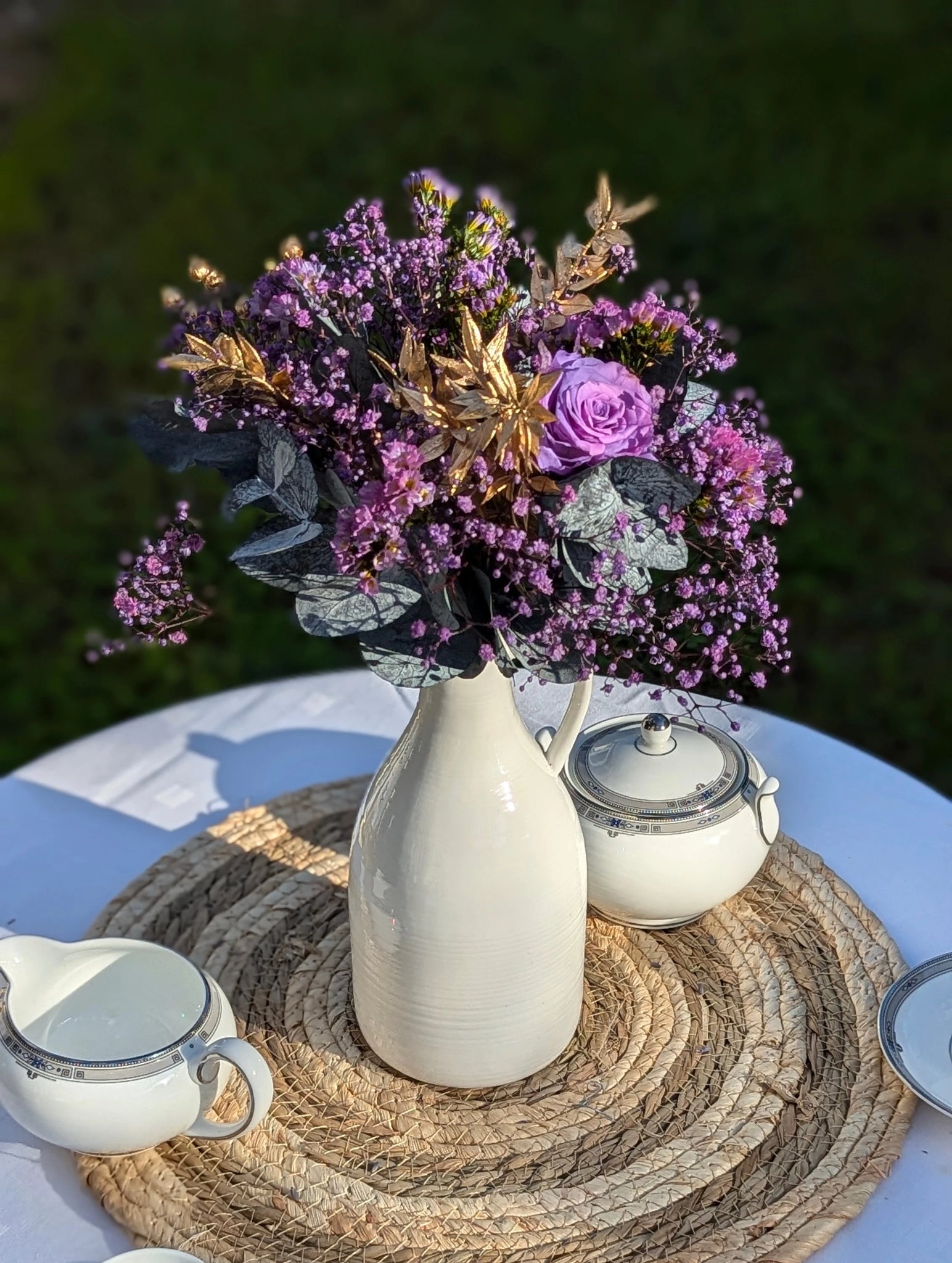 Bouquet violet et doré dans pichet blanc sur table en extérieur
