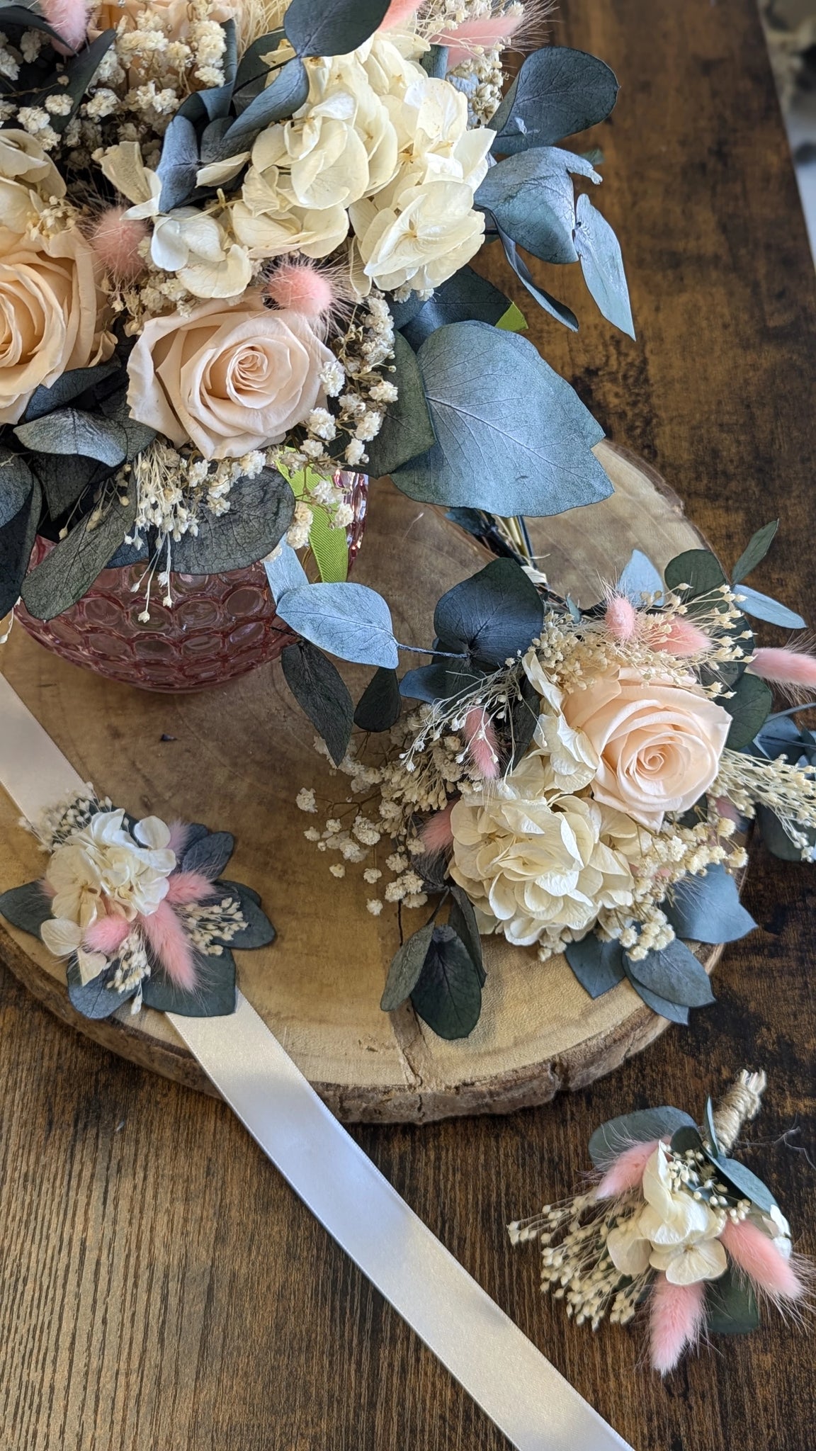 Iris. Bouquet de mariée en fleurs stabilisées composé de roses pastel, hortensias ivoire, eucalyptus bleuté, gypsophile et lagurus rose poudré, présenté dans un vase rose texturé, idéal pour un mariage romantique et durable. présenté avec ses accessoires