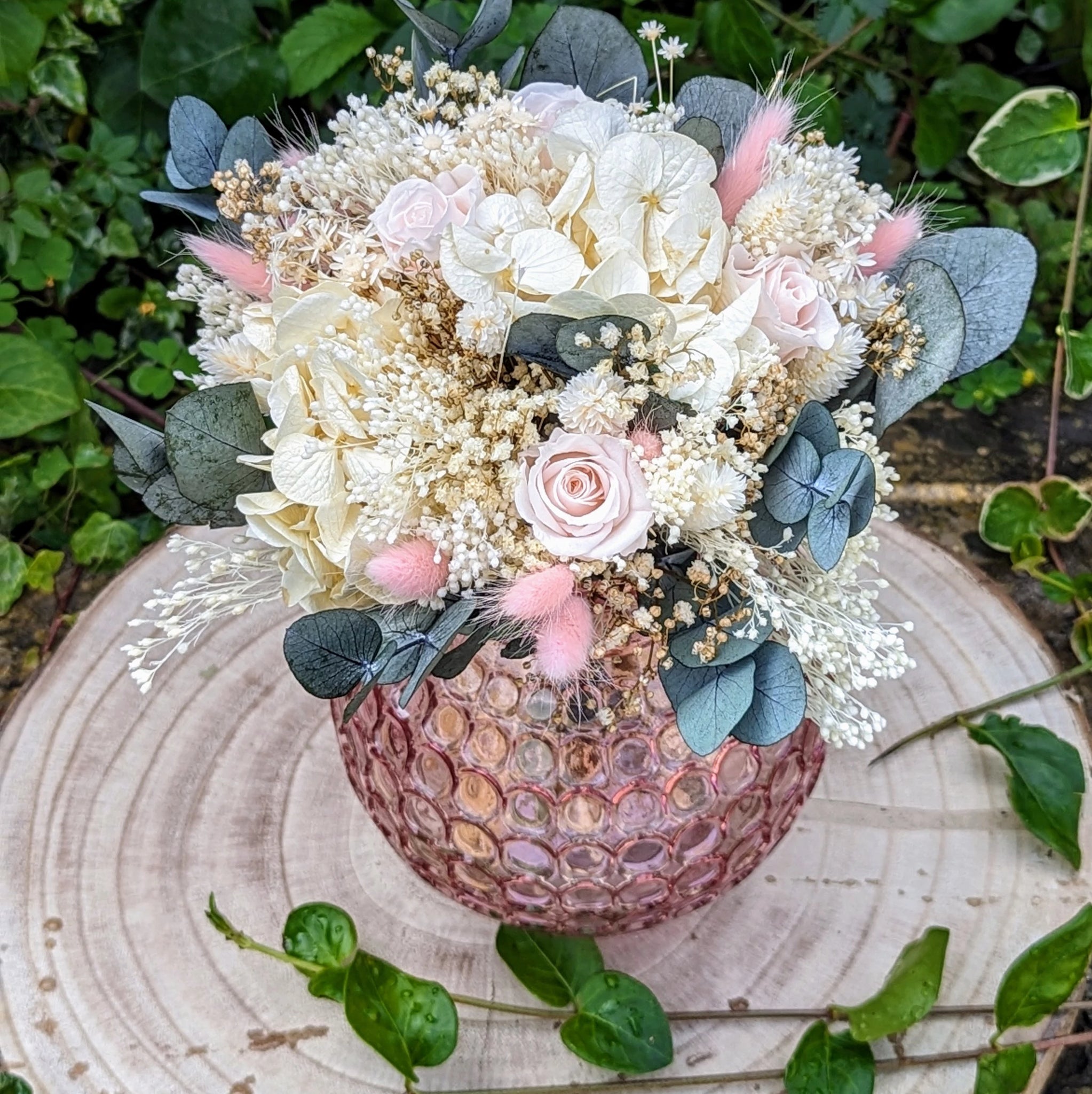 Bouquet de mariée en fleurs stabilisées composé de roses pastel, hortensias ivoire, eucalyptus bleuté, gypsophile et lagurus rose poudré, présenté dans un vase rose texturé, idéal pour un mariage romantique et durable.