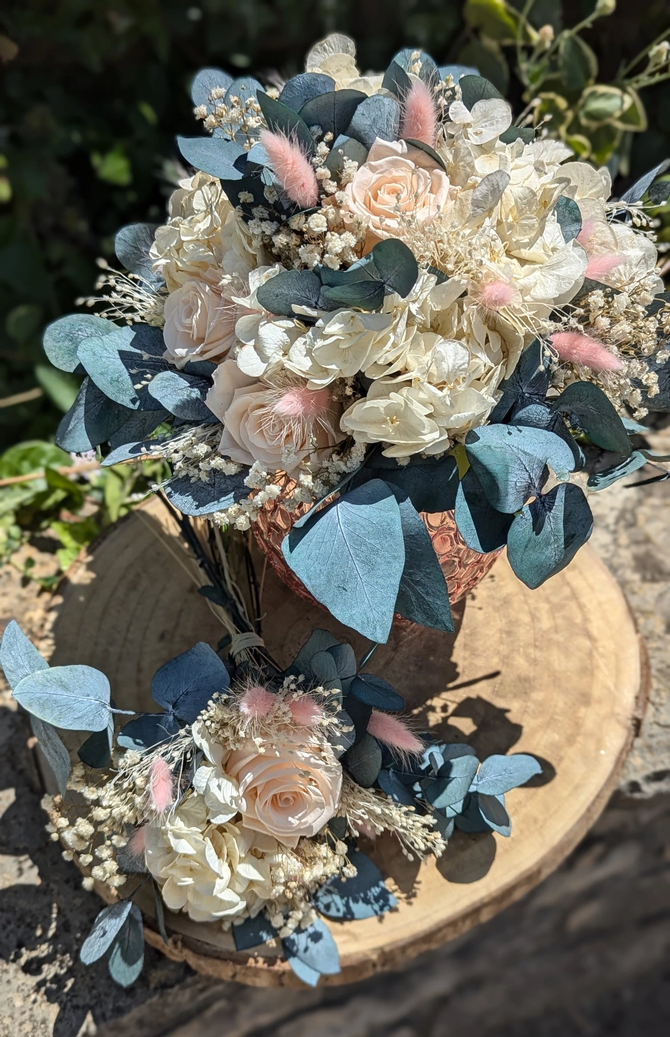 Iris. Bouquet de mariée en fleurs stabilisées composé de roses pastel, hortensias ivoire, eucalyptus bleuté, gypsophile et lagurus rose poudré, présenté dans un vase rose texturé, idéal pour un mariage romantique et durable. présenté en extérieur sur support de bois