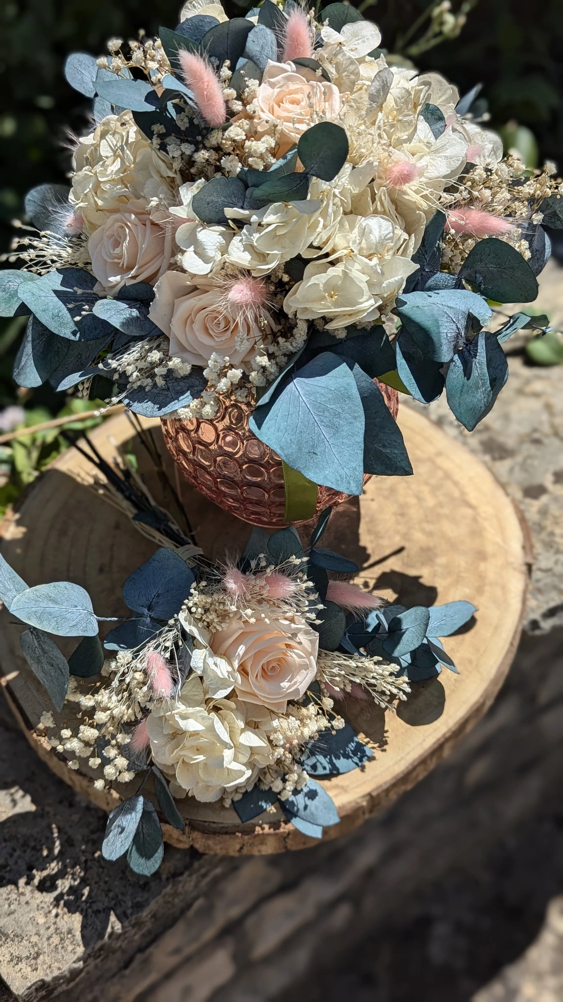 Iris. Bouquet de mariée en fleurs stabilisées composé de roses pastel, hortensias ivoire, eucalyptus bleuté, gypsophile et lagurus rose poudré, présenté dans un vase rose texturé, idéal pour un mariage romantique et durable. présenté en extérieur sur support de bois et le bouquet à jeter